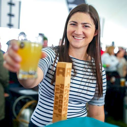 A woman is smiling and holding a glass of beer at an event, with people in the background. She is wearing a striped shirt and standing behind a counter.