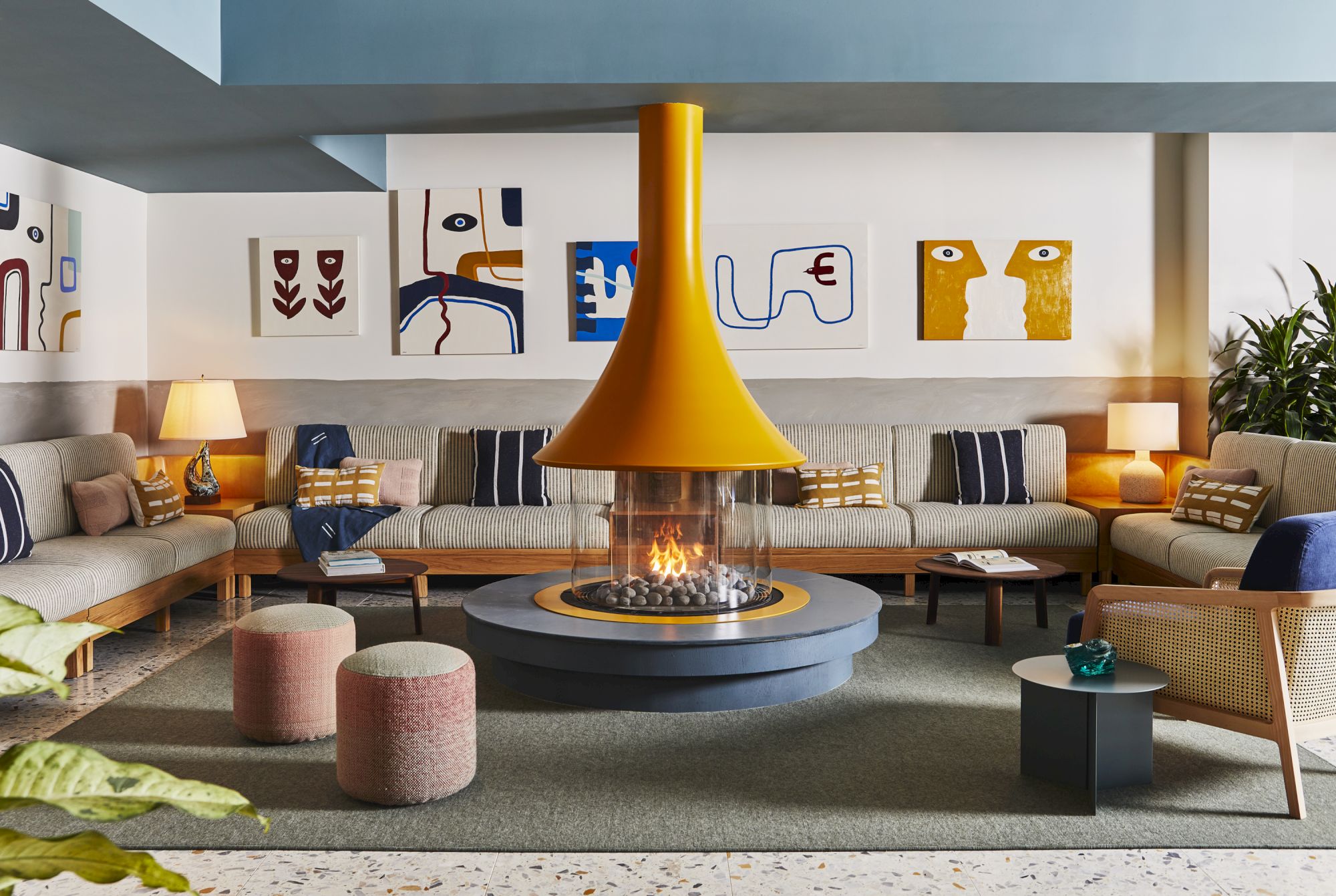 A modern living room with a central fireplace, surrounded by sofas, pillows, poufs, lamps, and abstract art on the walls.