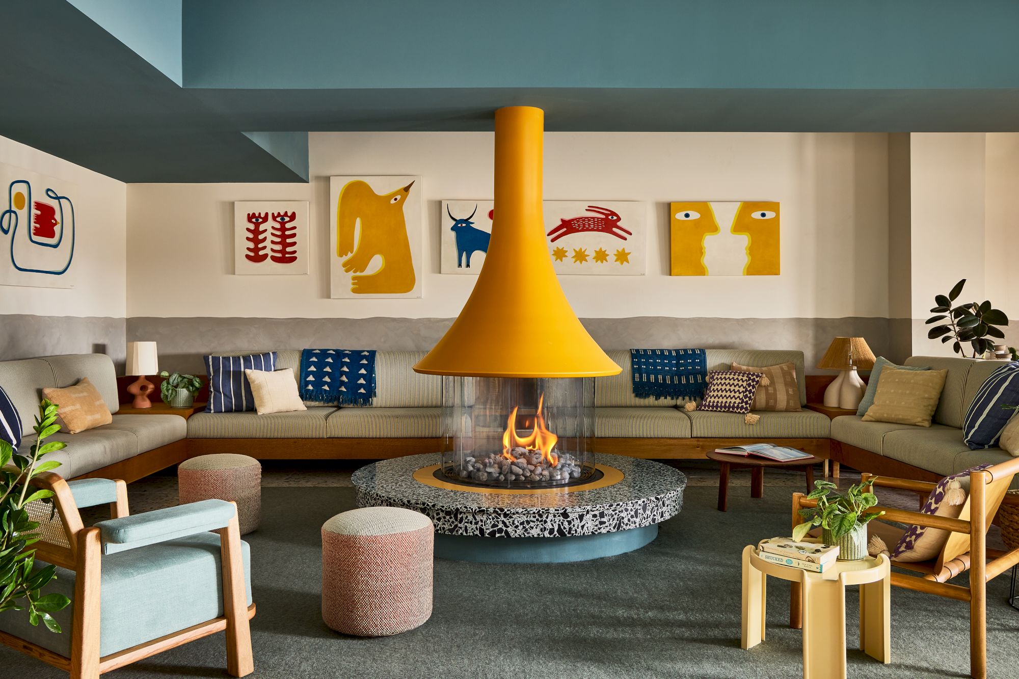 A cozy, modern lounge around a central fire pit with a bright yellow hanging centerpiece, circular stone hearth, and colorful abstract wall art, plus comfortable seating.