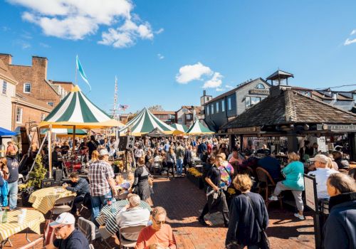 People enjoying an outdoor event with striped tents, tables, and chairs on a sunny day in a brick-paved area with historic-looking buildings around.