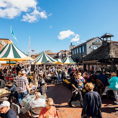 People enjoying an outdoor event with striped tents, tables, and chairs on a sunny day in a brick-paved area with historic-looking buildings around.