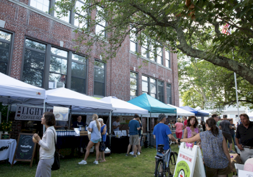 People are gathered at an outdoor market with various vendor tents set up in front of a brick building on a sunny day, engaging in activities.