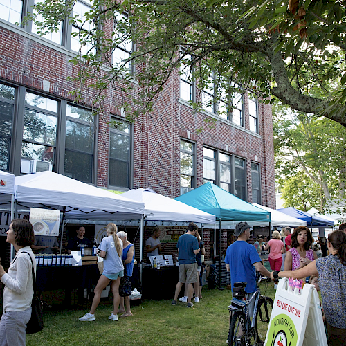 People are gathered at an outdoor market with various vendor tents set up in front of a brick building on a sunny day, engaging in activities.