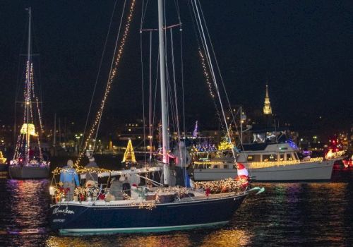 A nighttime boat parade with vessels decorated with festive lights on a waterway, set against a dark sky in a lit-up city.