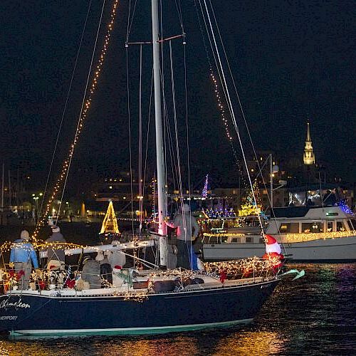 A nighttime boat parade with vessels decorated with festive lights on a waterway, set against a dark sky in a lit-up city.