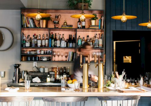 A cozy bar scene with shelves of liquor bottles, glasses, and decor, attended by a blurred bartender, with warm lighting and bar stools at the counter.