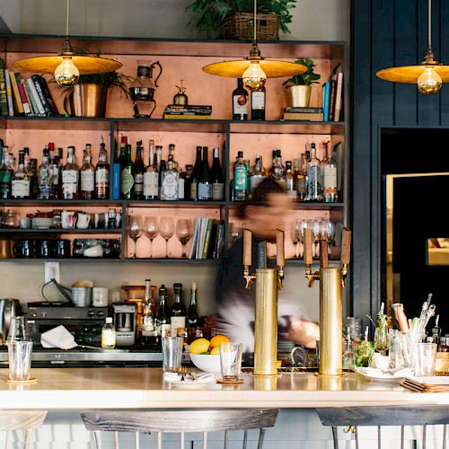 A cozy bar scene with shelves of liquor bottles, glasses, and decor, attended by a blurred bartender, with warm lighting and bar stools at the counter.