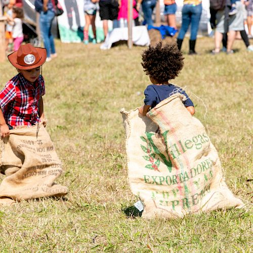 Two children are participating in a sack race on a grassy field, with one wearing a cowboy hat. Several people are gathering in the background.