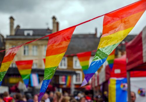 Colorful rainbow flags are strung up above a street filled with people, likely at a festive event, with buildings and other tents in the background.