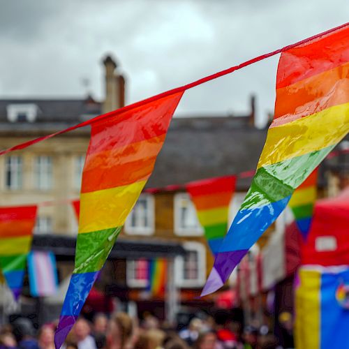 Colorful rainbow flags are strung up above a street filled with people, likely at a festive event, with buildings and other tents in the background.