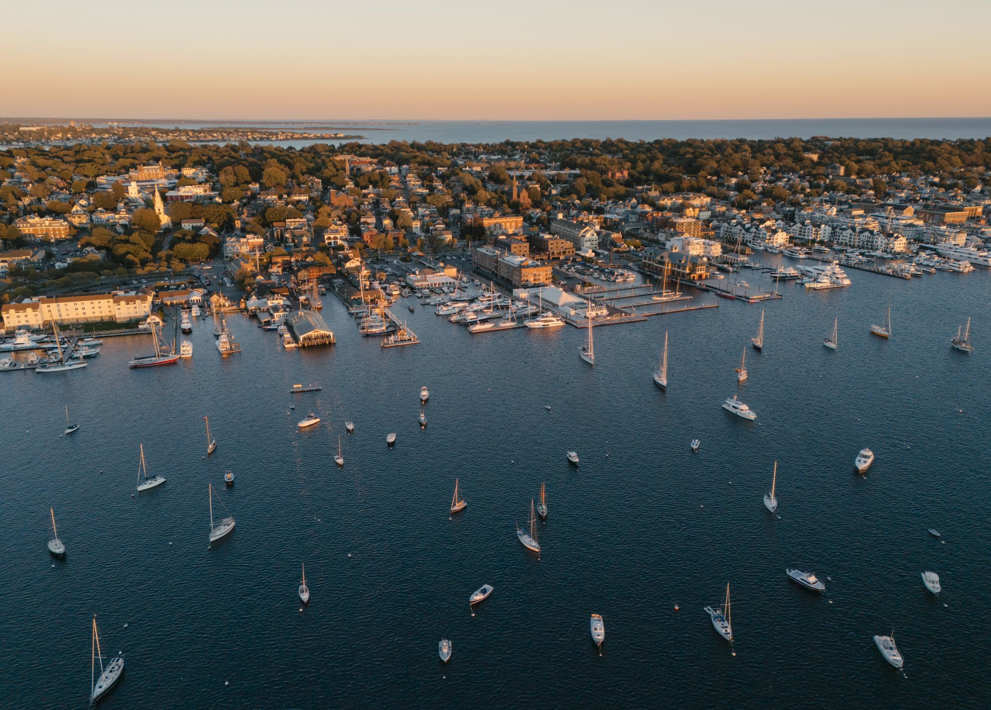 An aerial view of a coastal harbor packed with sailboats and yachts along the shoreline at sunset.