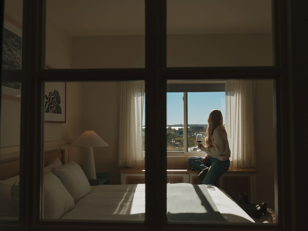 A woman sits by an open window holding a glass of wine, relaxing in a sunlit hotel room with a view outside.