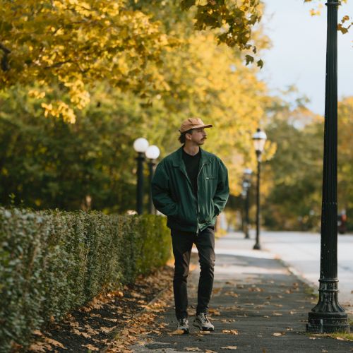 A man stands on an autumn sidewalk, hands in pockets, amid fallen leaves and golden trees with a streetlamp nearby.
