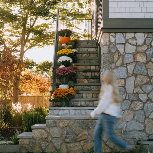 Sunny porch stairs with pumpkins and colorful flowers beside a stone wall, with a lantern and a person passing by blurred.