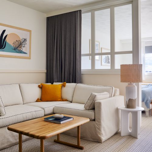 A bright, airy living room with a white sectional sofa, orange accent pillow, wooden coffee table, and framed wall art; a sliding glass door leads to a bedroom area.