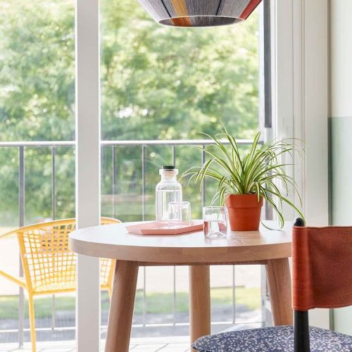 A bright dining nook with a light wood table, two chairs, a hanging geometric pendant, and a sunny window showing a wooden deck outside.