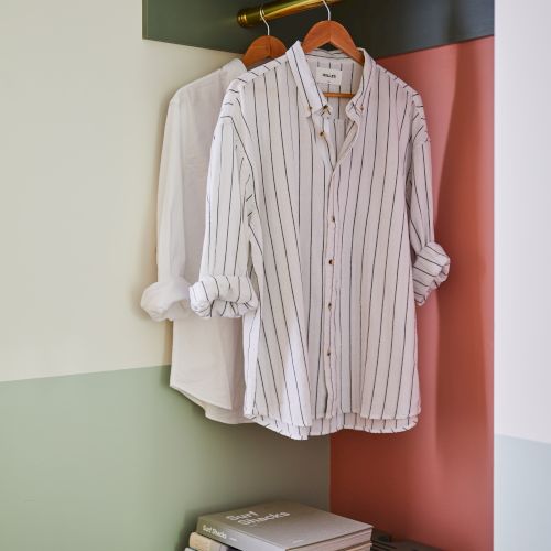 A striped shirt on a hanger in a closet, with folded clothes on a shelf and a hat nearby, all in a cozy wardrobe setup.