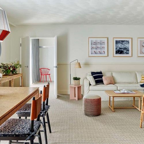 A bright, cozy living room with a small dining area, warm wood tones, a sofa and armchairs, wall art, and soft natural light from a window.