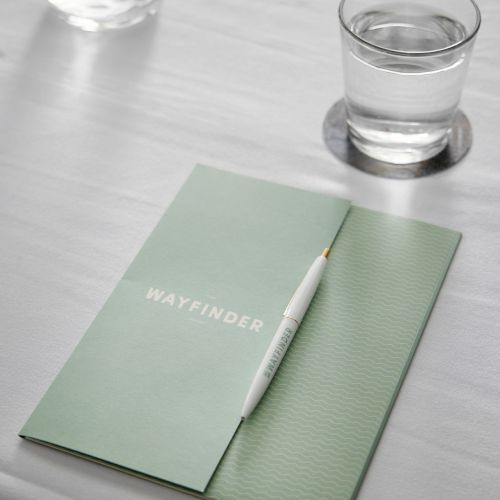A mint-green notebook with a white pencil resting on its pages, placed on a white table beside a glass of water and a bottle.