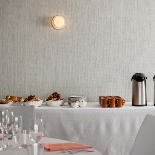 A breakfast or coffee setup on a white table with cups and two thermal carafes, empty plates and glasses, against a light wall.