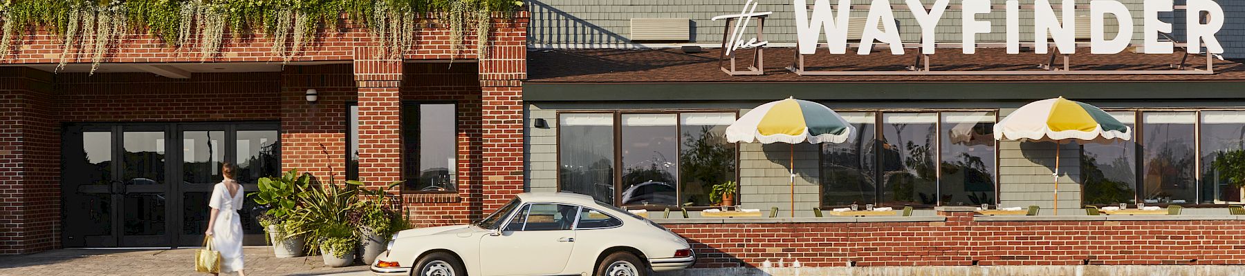 A vintage car parked in front of a building labeled "The Wayfinder," with a person walking by and colorful umbrellas above.