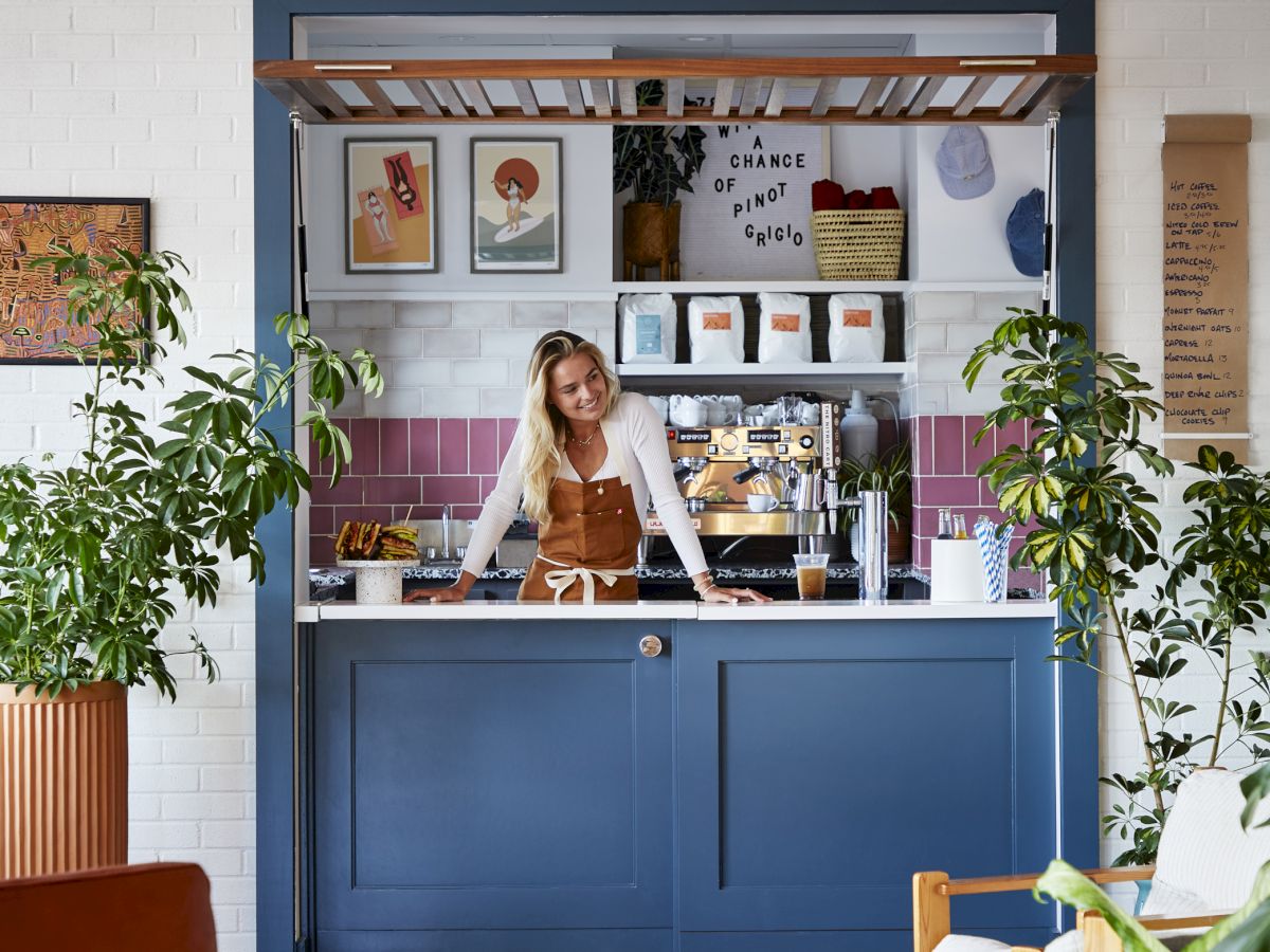 A cozy cafe with a blue counter, surrounded by plants and colorful decor, with a person behind the counter preparing coffee.