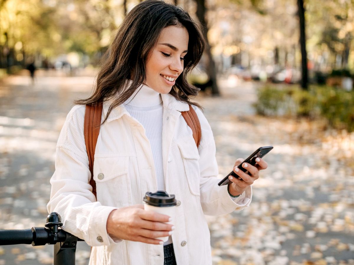 A person in a park uses their phone and holds a coffee cup while smiling. They are casually dressed and wearing a backpack during a fall day.