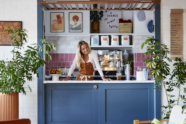 A woman stands behind the counter of a cozy coffee shop with blue cabinetry, surrounded by plants and colorful tiles, ready to serve customers.