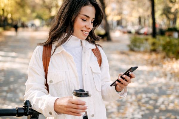 A person stands outdoors in a park, smiling while looking at their phone, holding a coffee cup, with autumn trees and fallen leaves around.