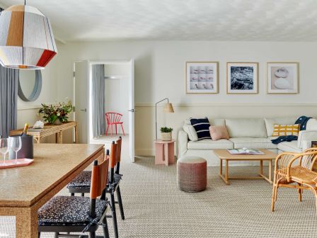 Modern living room with wooden dining table, colorful pendant light, white sofa, framed art, wicker chairs, and open door to another room.