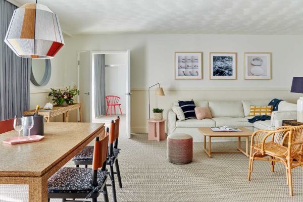 Modern living room with wooden dining table, colorful pendant light, white sofa, framed art, wicker chairs, and open door to another room.