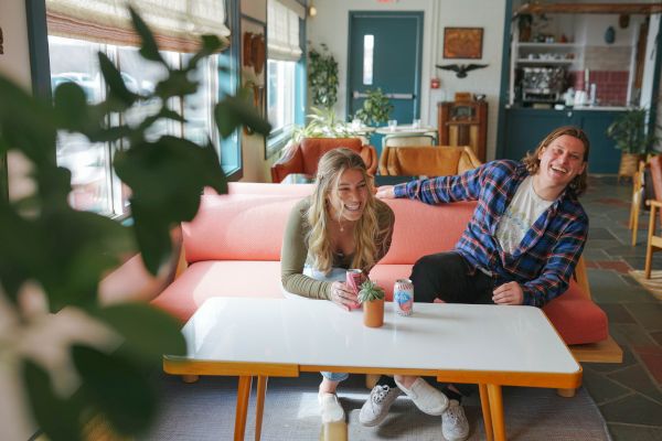 Two people are sitting on a pink couch in a cozy room, laughing and enjoying drinks at a white table.