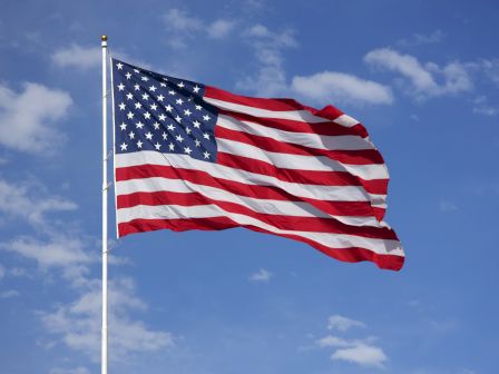 The image shows the flag of the United States of America waving in the wind against a blue sky with scattered clouds.