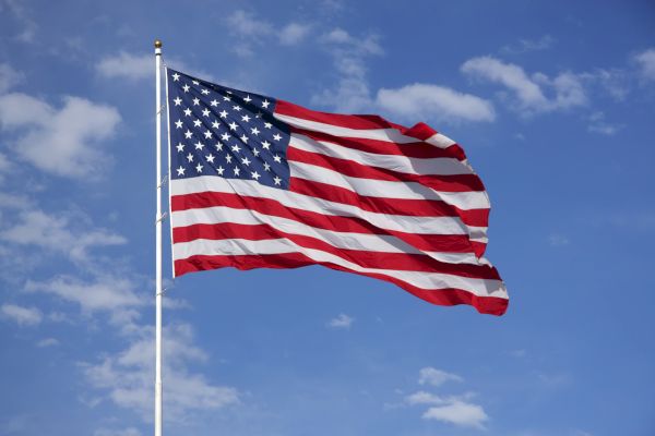 The image shows the flag of the United States of America waving in the wind against a blue sky with scattered clouds.