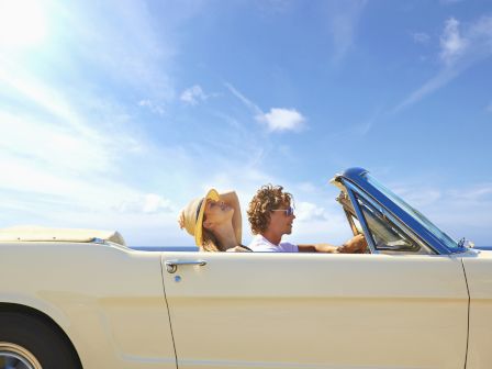 Two people are riding in a vintage convertible car with the top down under a clear blue sky. The passenger is leaning back, wearing a hat.