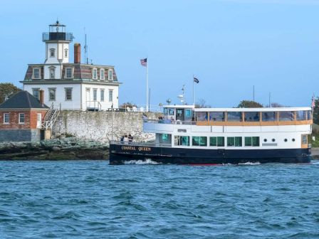 A lighthouse stands on a rocky shore with an American flag flying, and a passenger boat named Coastal Queen sails past in the foreground.