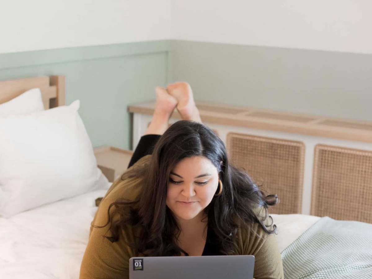 A person is lying on a bed, looking at a laptop, in a bright and cozy room.