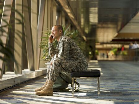 A person in military uniform is sitting on a bench inside a building, talking on a phone, with sunlight streaming through the windows.