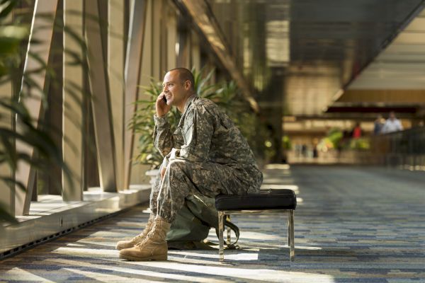 A person in military uniform is sitting on a bench inside a building, talking on a phone, with sunlight streaming through the windows.