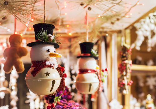 Hanging snowman ornaments with top hats and red scarves are surrounded by festive lights and decorations in a warm, cozy setting.