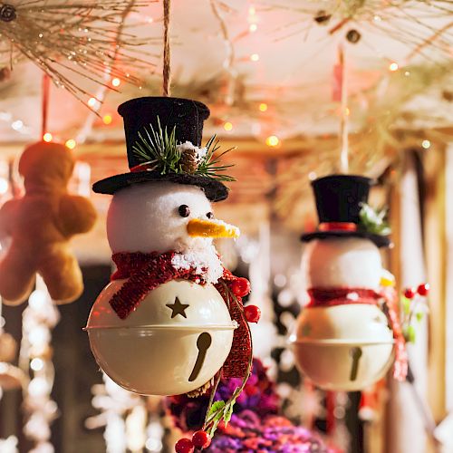 Hanging snowman ornaments with top hats and red scarves are surrounded by festive lights and decorations in a warm, cozy setting.