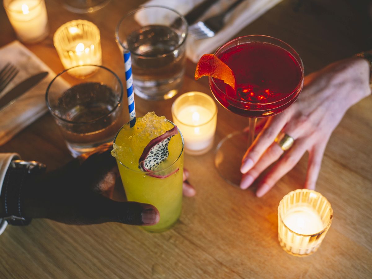 Two people hold drinks at a candlelit table. One glass is yellow with a dragon fruit garnish, the other is a red cocktail.