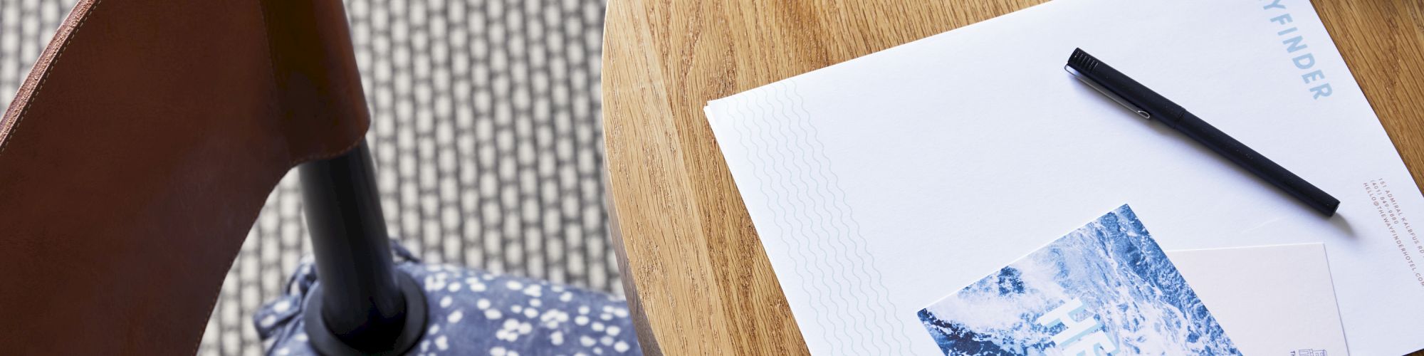 A table with a notebook, pen, postcard, sunglasses, and a patterned book, next to a chair with a blue and white polka dot cushion.