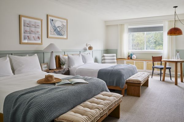 The image shows a cozy bedroom with two beds, a table, chairs, and wall art. Natural light streams through a window, adding warmth.