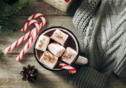 A cozy scene with hot chocolate topped with marshmallows and cocoa, surrounded by candy canes, a gray sweater, and a pinecone on wood.