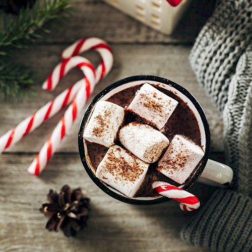 A cozy scene with hot chocolate topped with marshmallows and cocoa, surrounded by candy canes, a gray sweater, and a pinecone on wood.