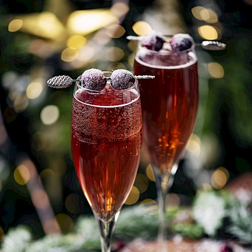 Two glasses of red sparkling drink, garnished with berries on skewers, surrounded by greenery and bokeh lights in the background.