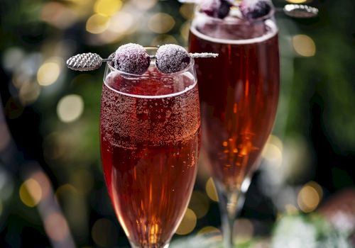 Two glasses of red sparkling drink, garnished with berries on skewers, surrounded by greenery and bokeh lights in the background.