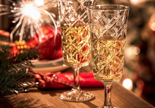 Two elegant crystal glasses filled with a festive drink are on a table, with a sparkling light and Christmas tree in the background.