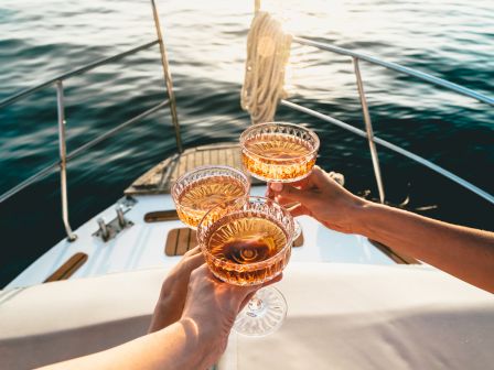 Three hands holding glasses of rosé wine, toasting on the deck of a boat with a scenic ocean view in the background.